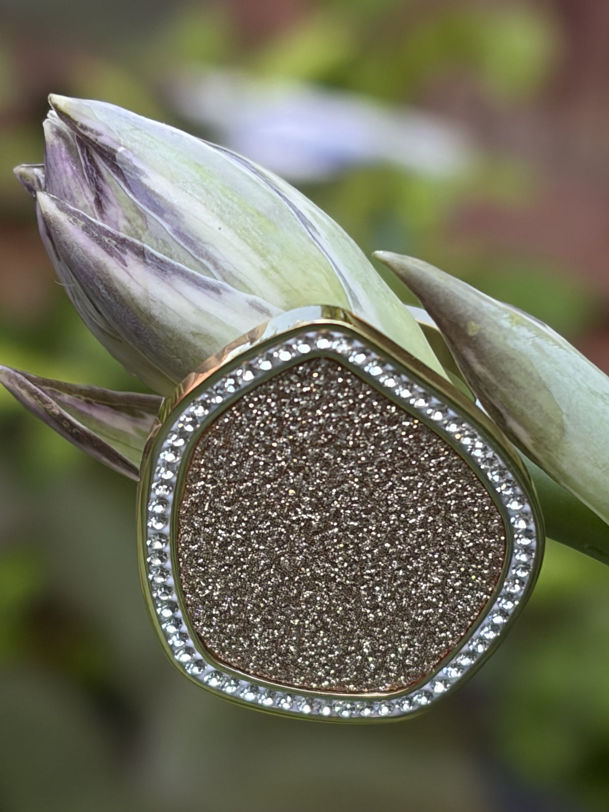 A large statement ring with a textured metallic gold center and a border of clear stones, displayed on a green lotus bud against a blurred natural background.